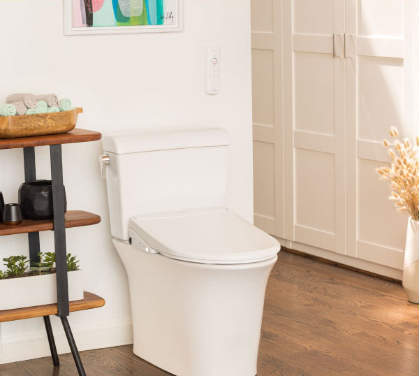 A sleek bathroom features the white 'Thinline' toilet seat, wooden shelves with towels and toiletries, a white cabinet, and a framed abstract painting on the wall.