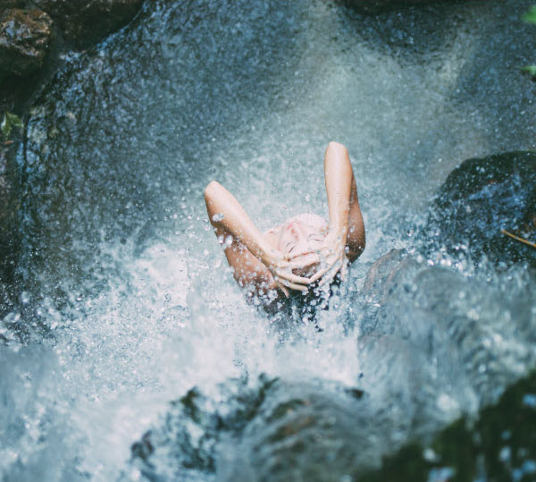 Person standing under a waterfall with arms raised, surrounded by splashing water, capturing a moment of refreshment and exhilaration.