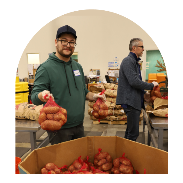 Two people at a food bank holding bags of potatoes, standing near tables filled with with supplies in an indoor setting. 