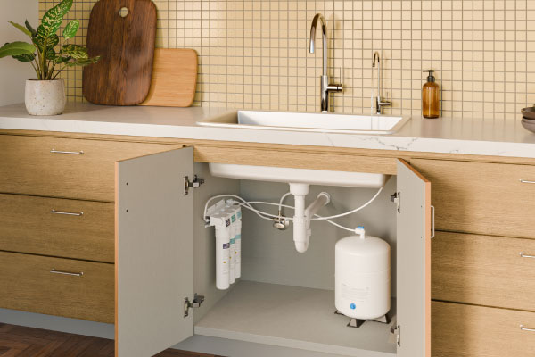 Modern kitchen sink with open cabinet showing under-sink water filtration system. Countertop has a plant, cutting boards, and soap dispenser.