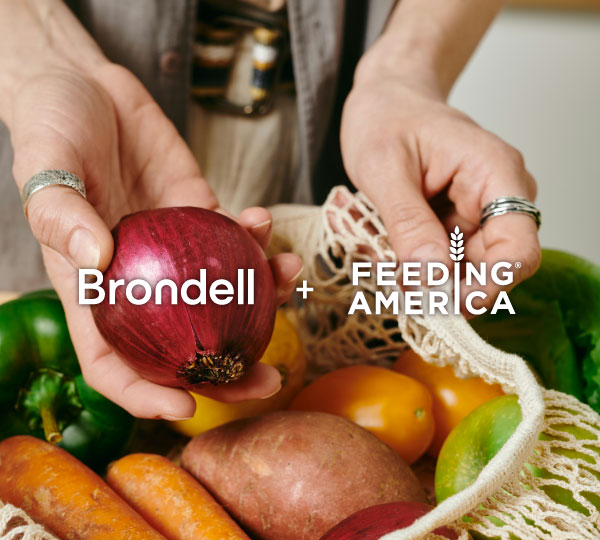 Person holding a red onion above a reusable bag filled with carrots, bell peppers, and potatoes. Text overlay reads 'Brondell + Feeding America'.