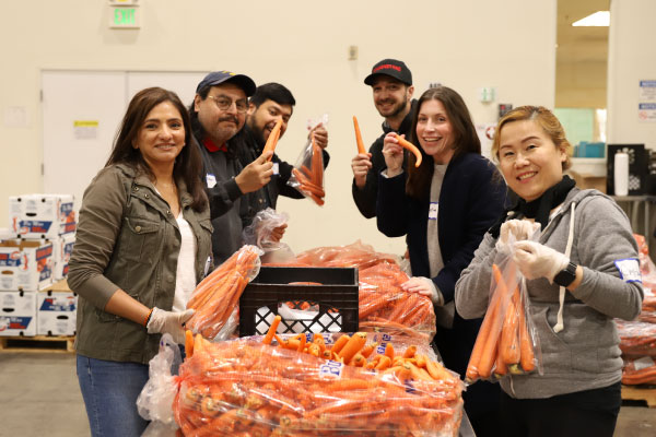 Brondell employees at a food bank packaging carrots at a table. They wear gloves and name tags, with boxes stacked in the background of the distribution area.