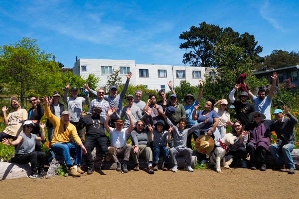 Group of about 25 people posing outdoors in a garden or park with trees, bushes, and a building in the background. A dog sits in front; some raise their hands.