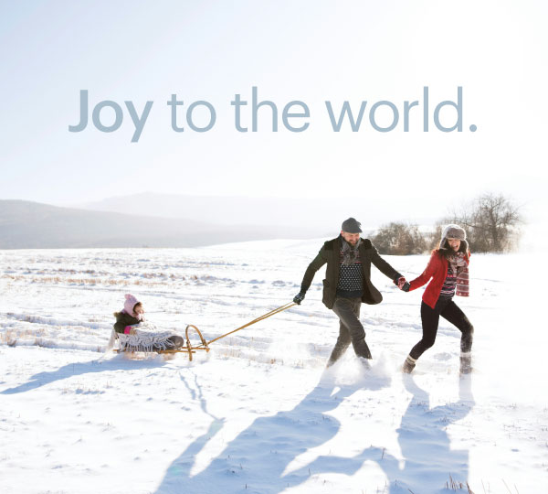 "Family enjoying a snowy day as two adults run hand-in-hand across a snow-covered field while pulling a child on a wooden sled, with bright sunlight and rolling hills in the background.