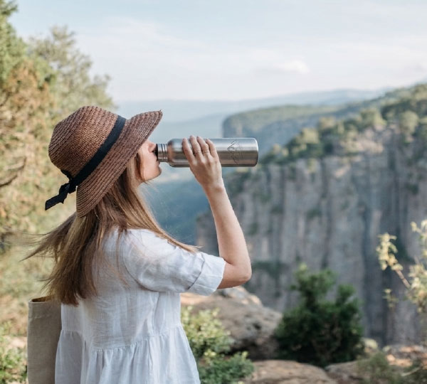 Person wearing a sun hat drinks from a reusable water bottle while overlooking a scenic mountain landscape, highlighting clean hydration and sustainable living outdoors.